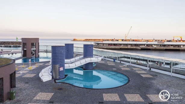 A rooftop pool area with a blue-tiled structure, a small building, and a view of the sea and port in the background. - Home Azores - Mountain View Apartment (Ponta Delgada)