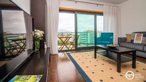 A modern living room with a flat-screen TV, a sofa, a coffee table, and a balcony with a view of buildings. - Home Azores - Mountain View Apartment (Ponta Delgada)