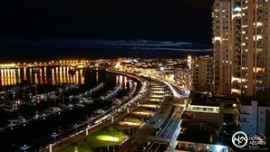 A nighttime aerial view of a coastal city with a marina, illuminated buildings, and a prominent high-rise hotel. - Home Azores - Mountain View Apartment (Ponta Delgada)