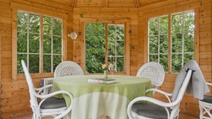 Uma cabana de madeira com uma mesa redonda e quatro cadeiras de vime branco, cercada por amplas janelas.