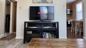 A living room with a TV displaying 'NETFLIX', a black entertainment unit with books, a wooden coffee table, and a kitchen visible in the background.