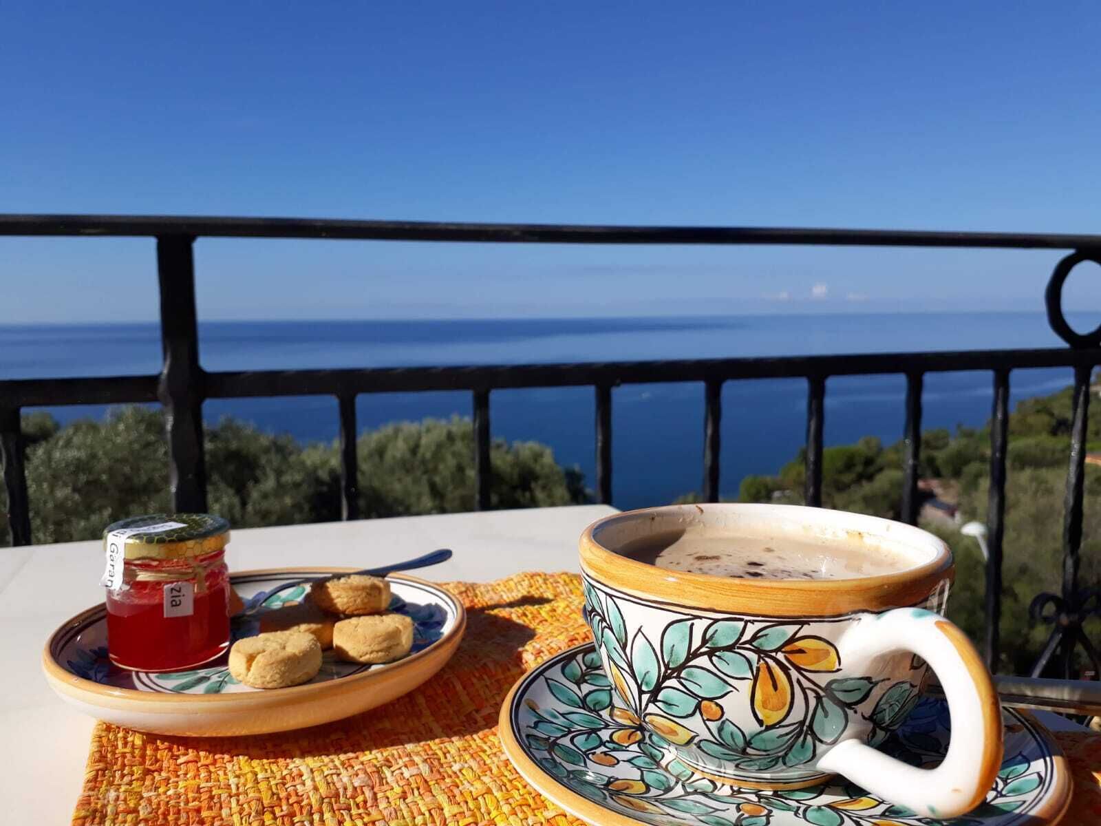 A table with a cup and saucer, a plate with cookies, and a jar of jam on a balcony overlooking the sea.