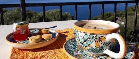 A table with a cup and saucer, a plate with cookies, and a jar of jam on a balcony overlooking the sea.