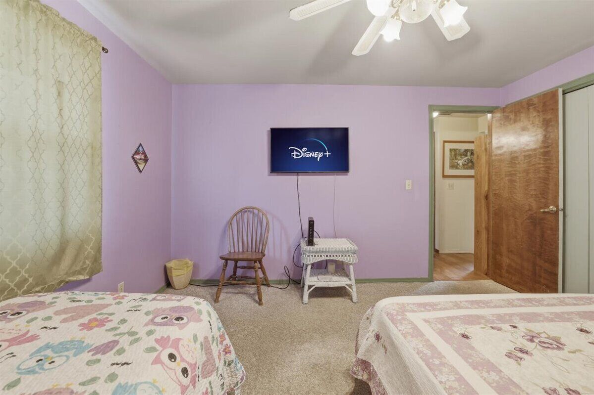 Un dormitorio con dos camas, una silla, una mesita, un televisor montado en la pared y una puerta que da a otra habitación.