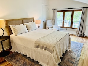 A bedroom with a large bed, a chair, a window with curtains, and a lamp on a bedside table.