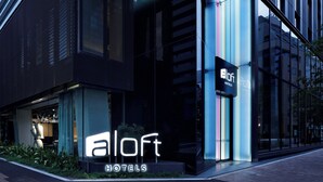 A modern hotel exterior with a prominent 'aLoft Hotels' sign, large glass windows, and a well-lit entrance.