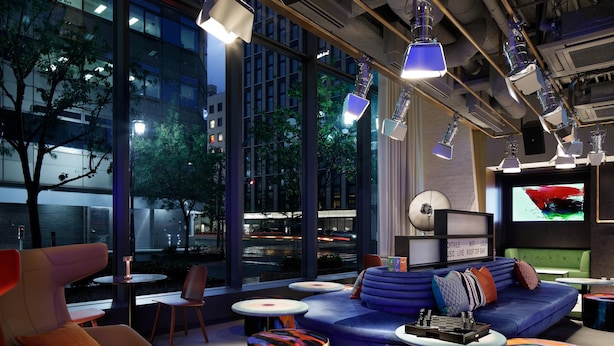 A modern lounge area with a sofa, chairs, and round tables. Large windows offer a view of the cityscape.