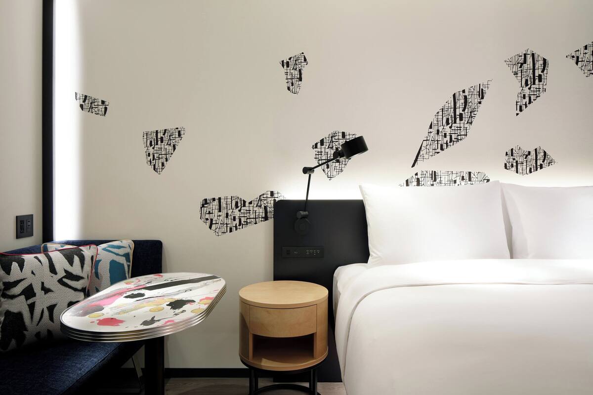 A hotel room with a bed, a bedside table with a lamp, a wall with abstract leaf decals, and a patterned pillow on the couch.