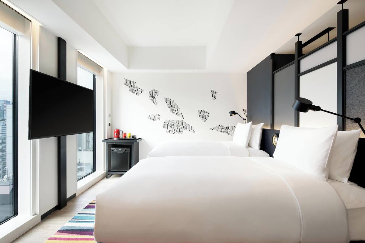 A modern hotel room with a large bed, a flat-screen TV mounted on the wall, a desk with a lamp, and a city view through large windows.