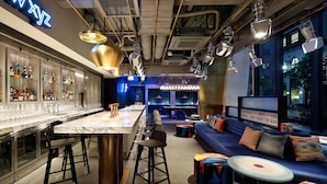 A bar area with a marble countertop, stools, and a neon sign. There is a seating area with a sofa and cushions, and a bar counter with a sign indicating available services.