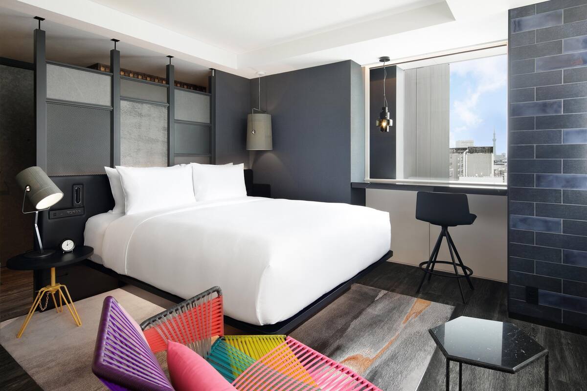 A modern hotel room with a large bed, a small table, a chair, and a view of the city through a large window.