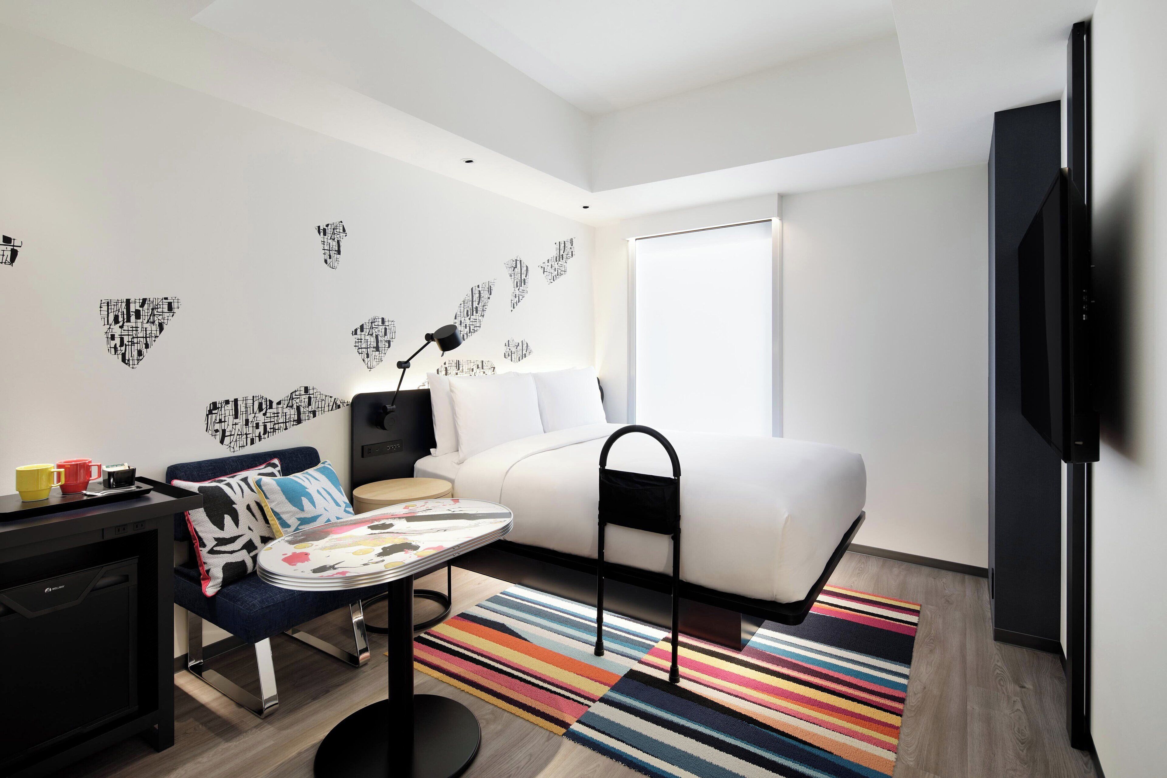Una habitación de hotel moderna con un sofá blanco, una mesita con una bandeja de objetos, una alfombra colorida a rayas y una pared con arte abstracto.