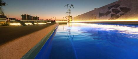 A swimming pool with a geometric wall design, a palm tree, and a building in the background.