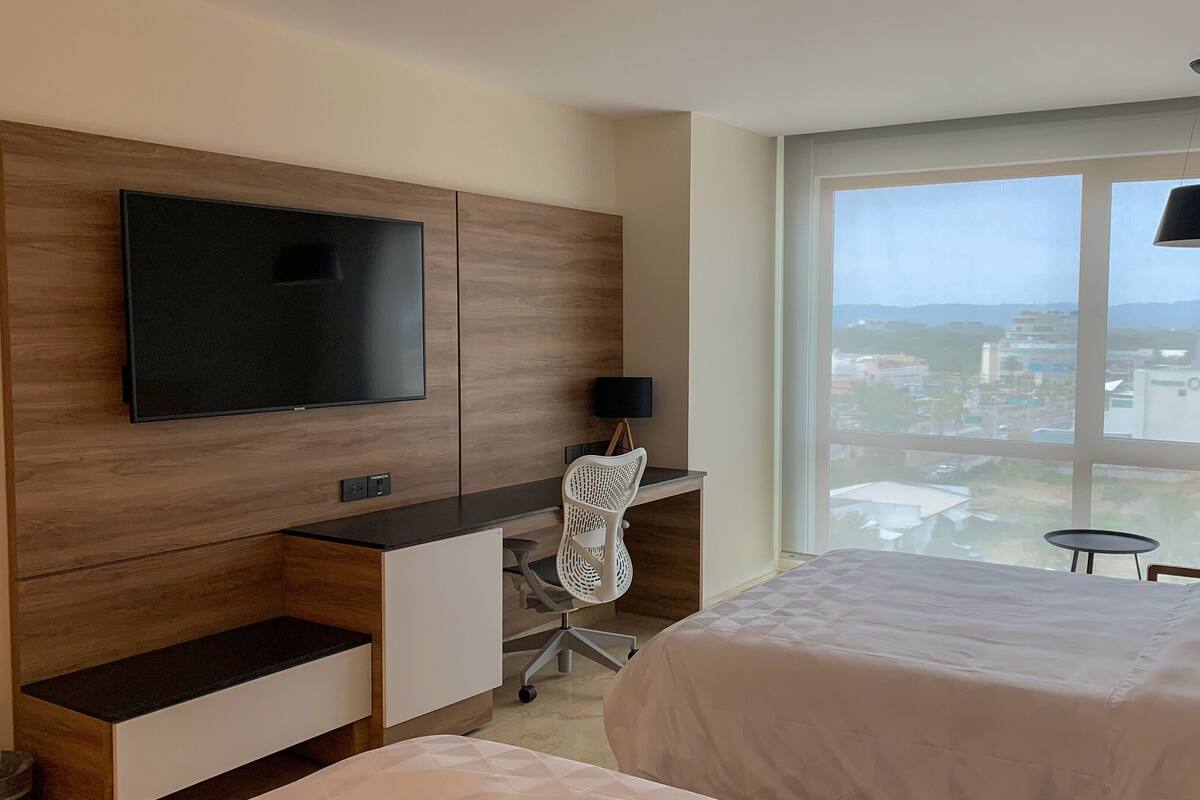 Une chambre d’hôtel moderne dotée d’une grande fenêtre donnant sur la ville, d’un téléviseur à écran plat fixé à un meuble en bois, d’un bureau avec une chaise et d’un lit recouvert d’une couette blanche.