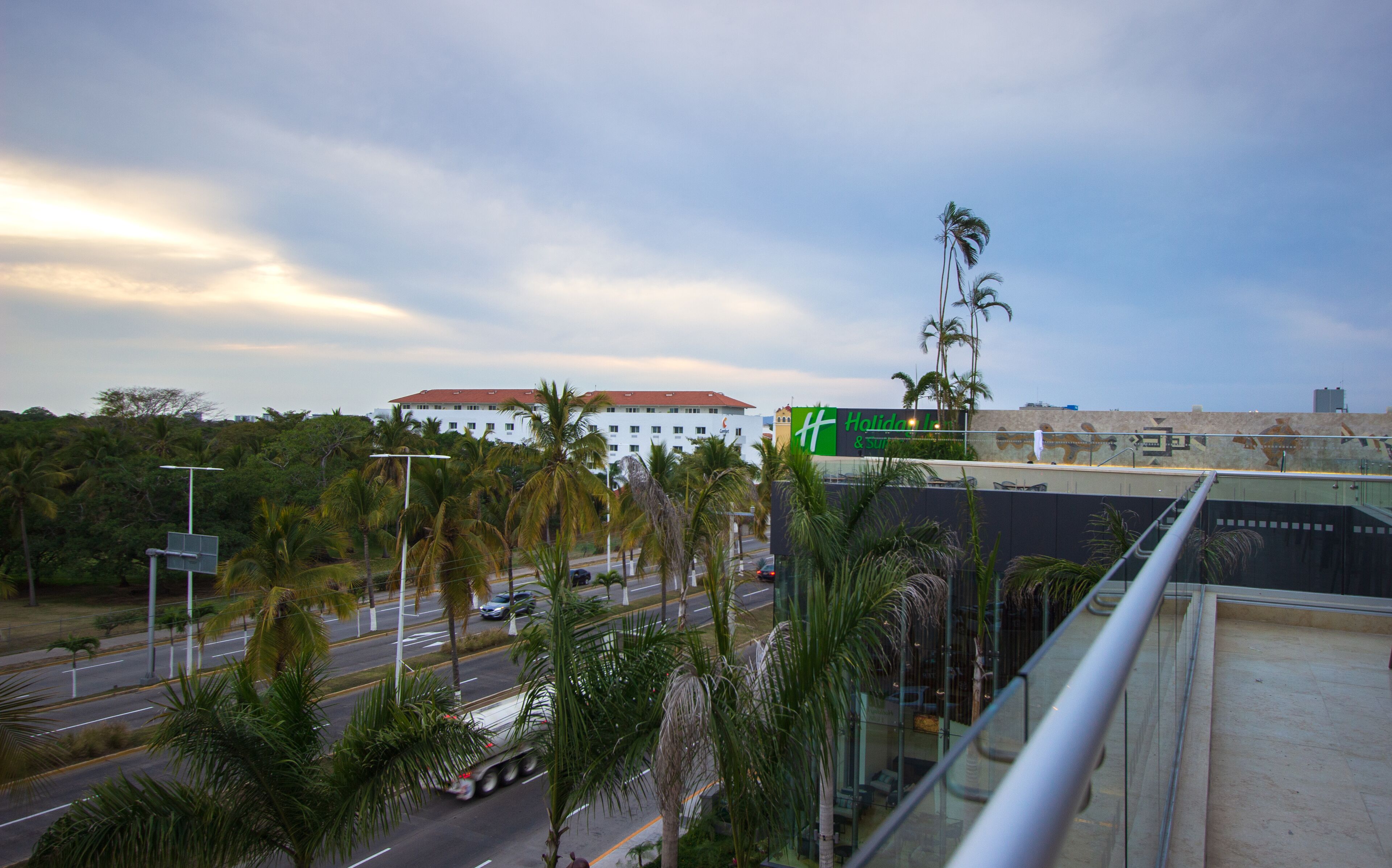 A view of a road with palm trees, a large building with a red roof, and a billboard advertising a hotel.