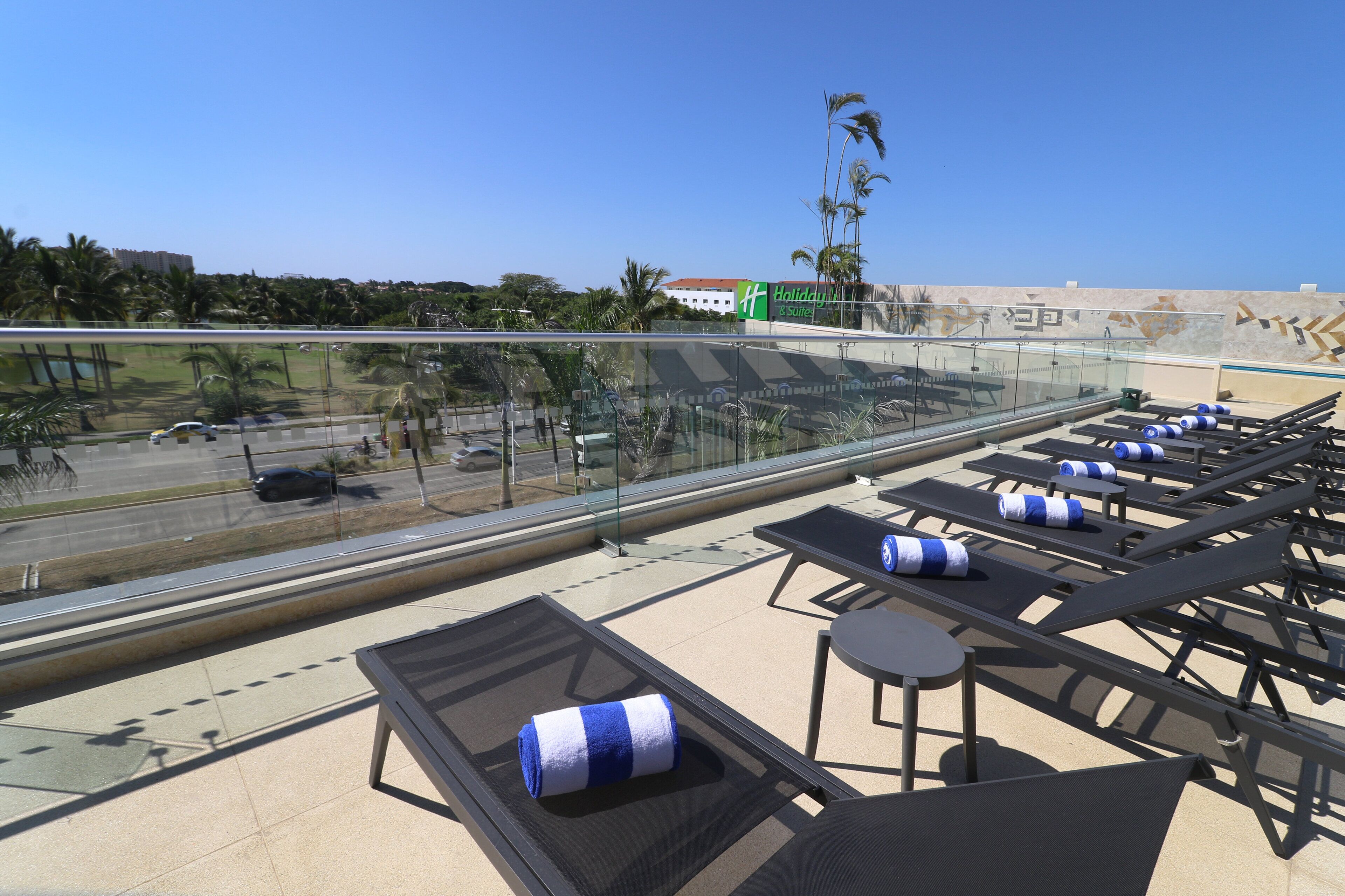 A rooftop lounge area with sun loungers, a glass balustrade, and a view of the road and buildings in the distance.