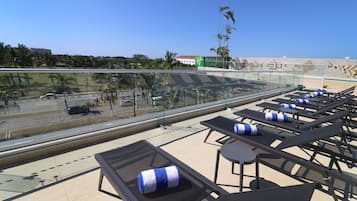 Un área de lounge en la azotea con sillones reclinables, barandilla de cristal y vistas a la calle y edificios a lo lejos.