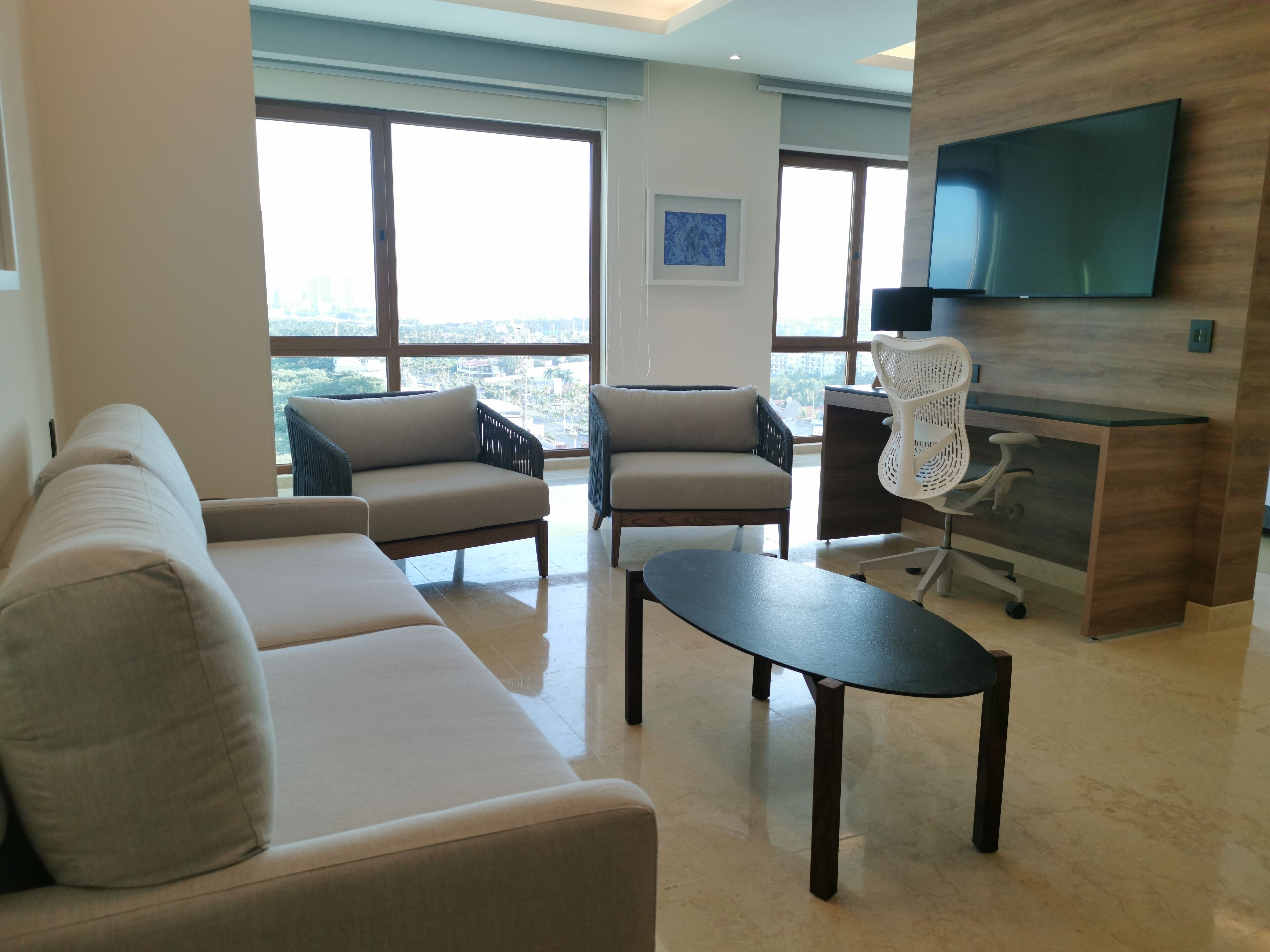 A modern living room with a sofa, two armchairs, a coffee table, and a desk with a chair. A flat-screen TV is mounted on the wall.