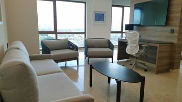 A modern living room with a sofa, two armchairs, a coffee table, and a desk with a chair. A flat-screen TV is mounted on the wall.