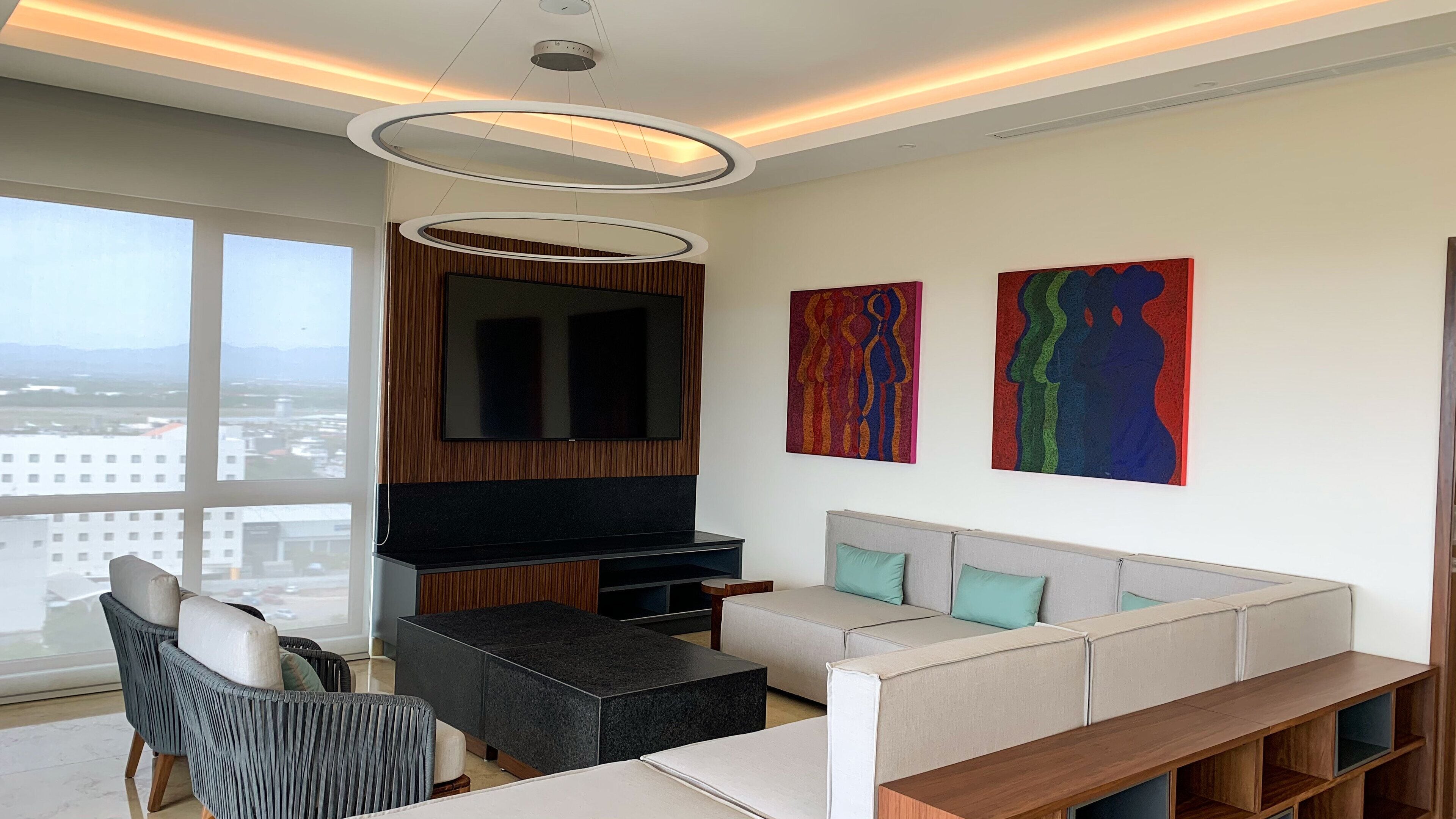 A modern living room with a large TV, a sofa, a coffee table, and a fireplace. There are two abstract paintings on the wall and a large window offering a city view.