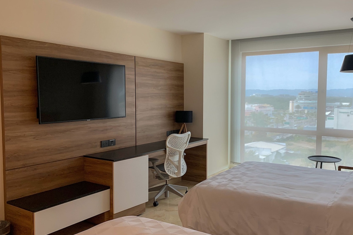 A hotel room with a bed, a desk with a chair, a TV mounted on the wall, and a large window with a view of the city.