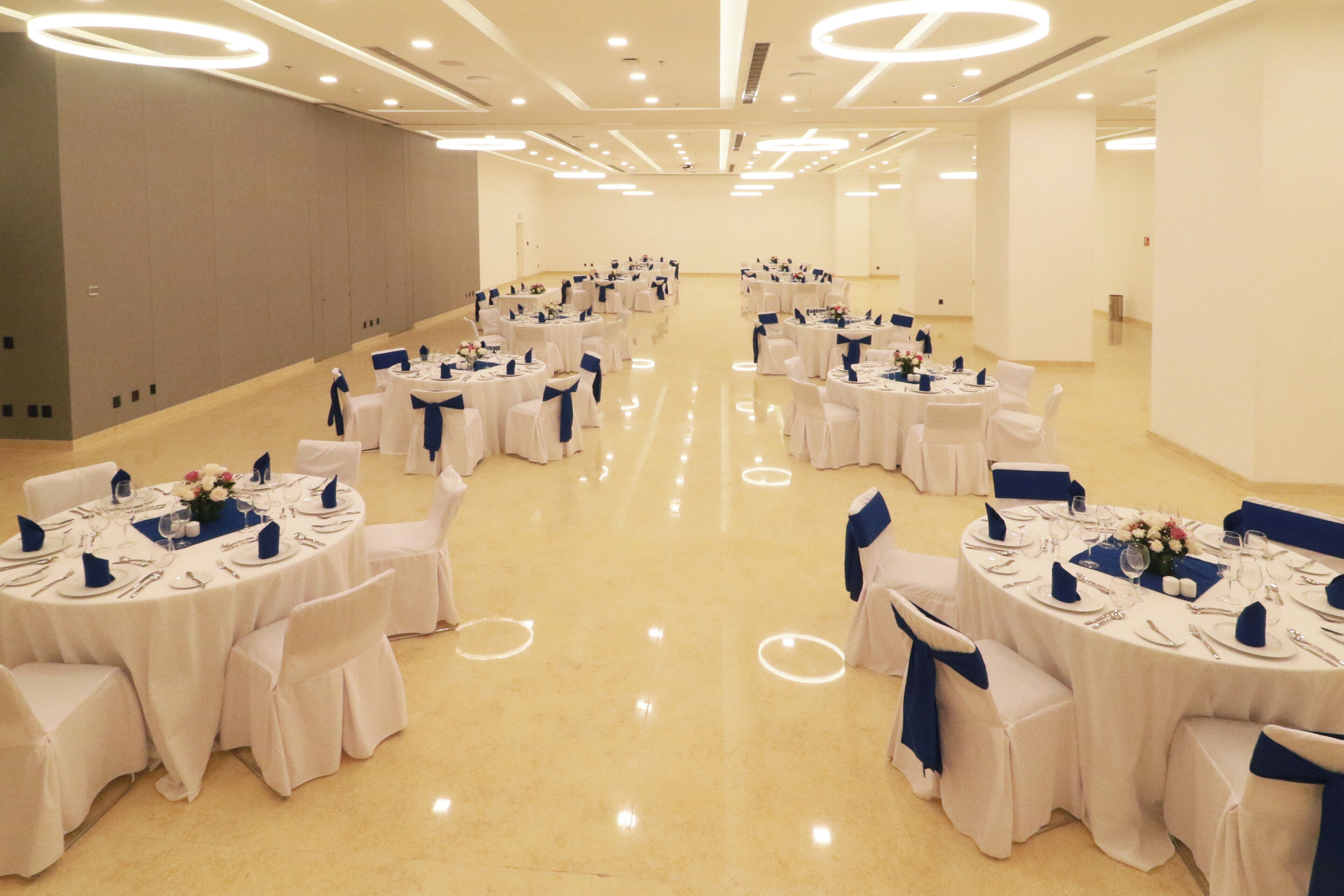 A large, well-lit banquet hall with multiple round tables set for dining, each with white tablecloths and blue chair covers.