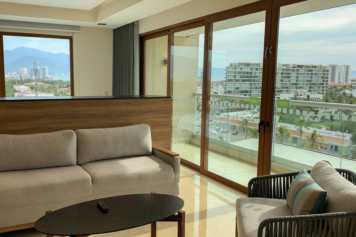 Un salon moderne avec un canapé, une table basse et une vue sur le paysage urbain, grâce à de grandes fenêtres.