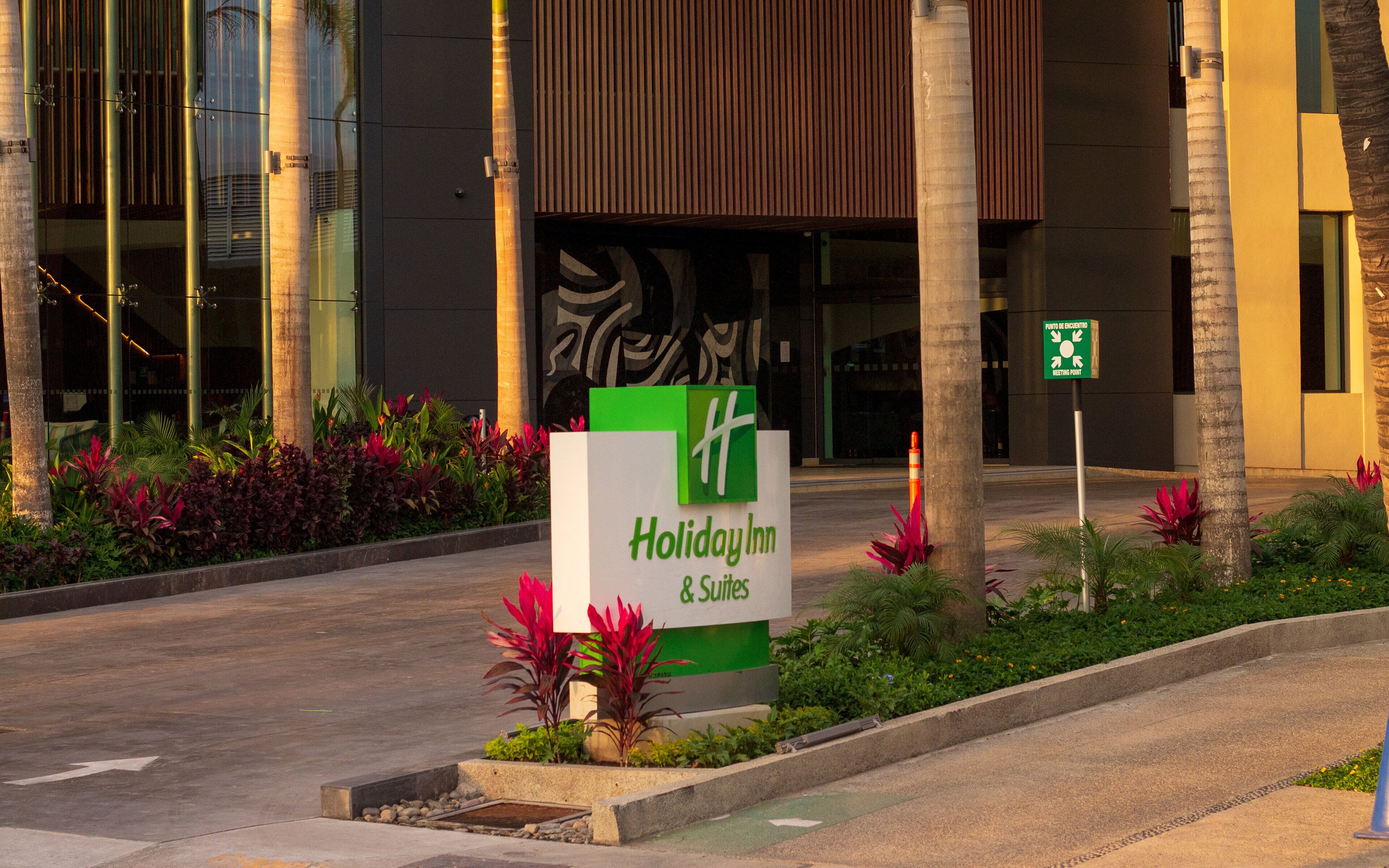 A Holiday Inn & Suites sign in front of a building with a modern design, palm trees, and a landscaped garden.