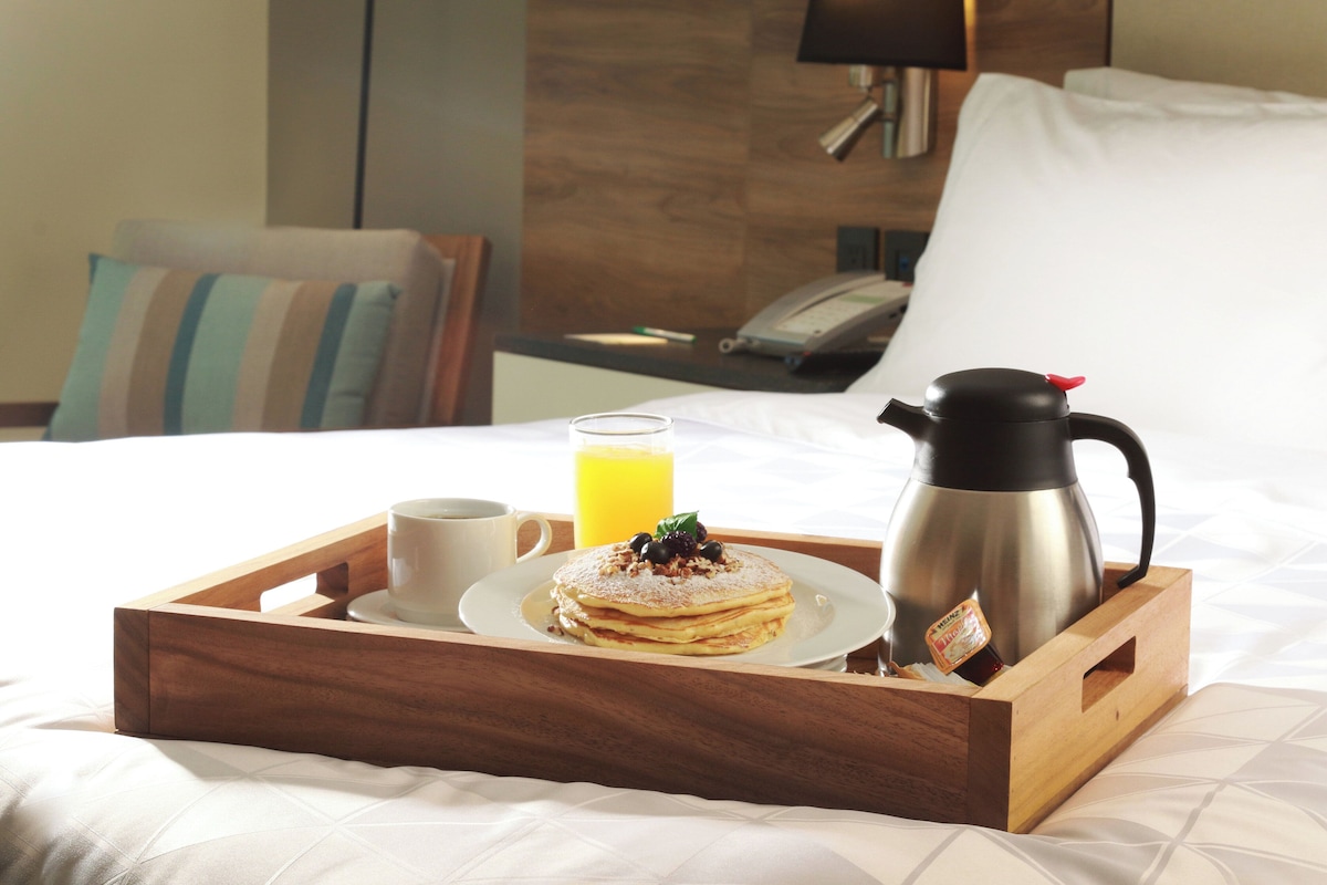 A breakfast tray with pancakes, a coffee cup, a glass of orange juice, and a coffee pot on a bed.