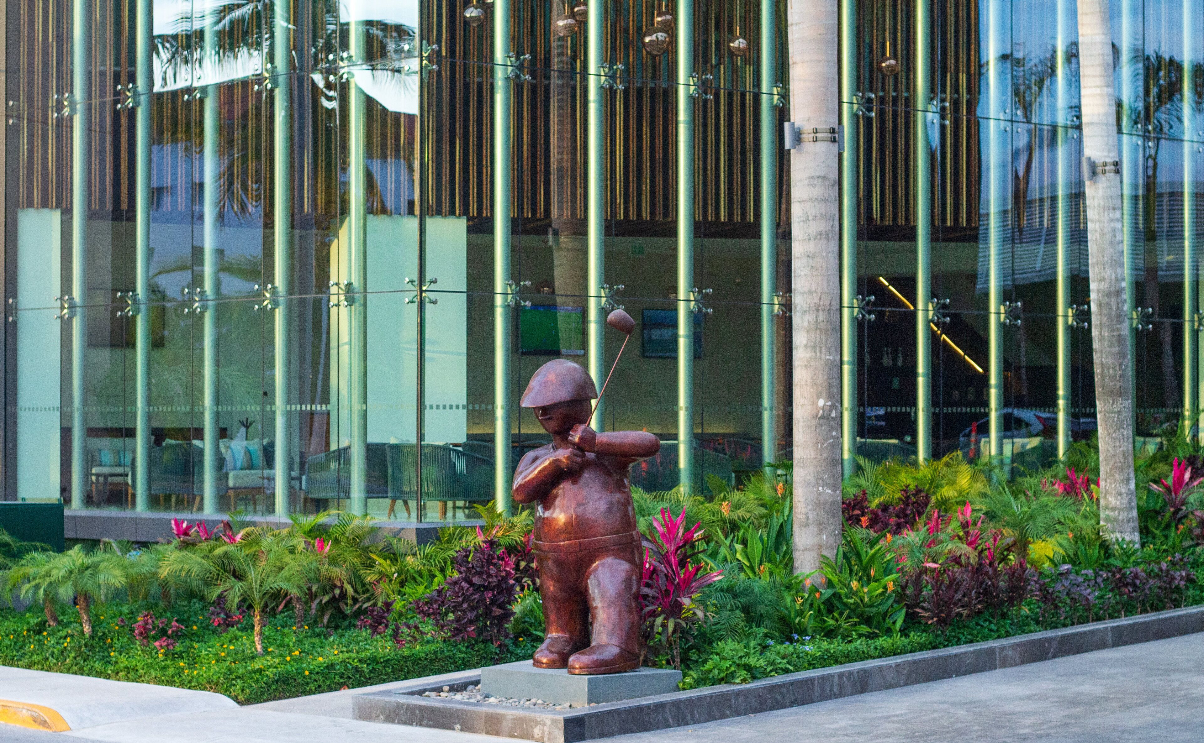 A bronze statue of a soldier holding a rifle in front of a modern building with vertical glass windows and tropical landscaping.