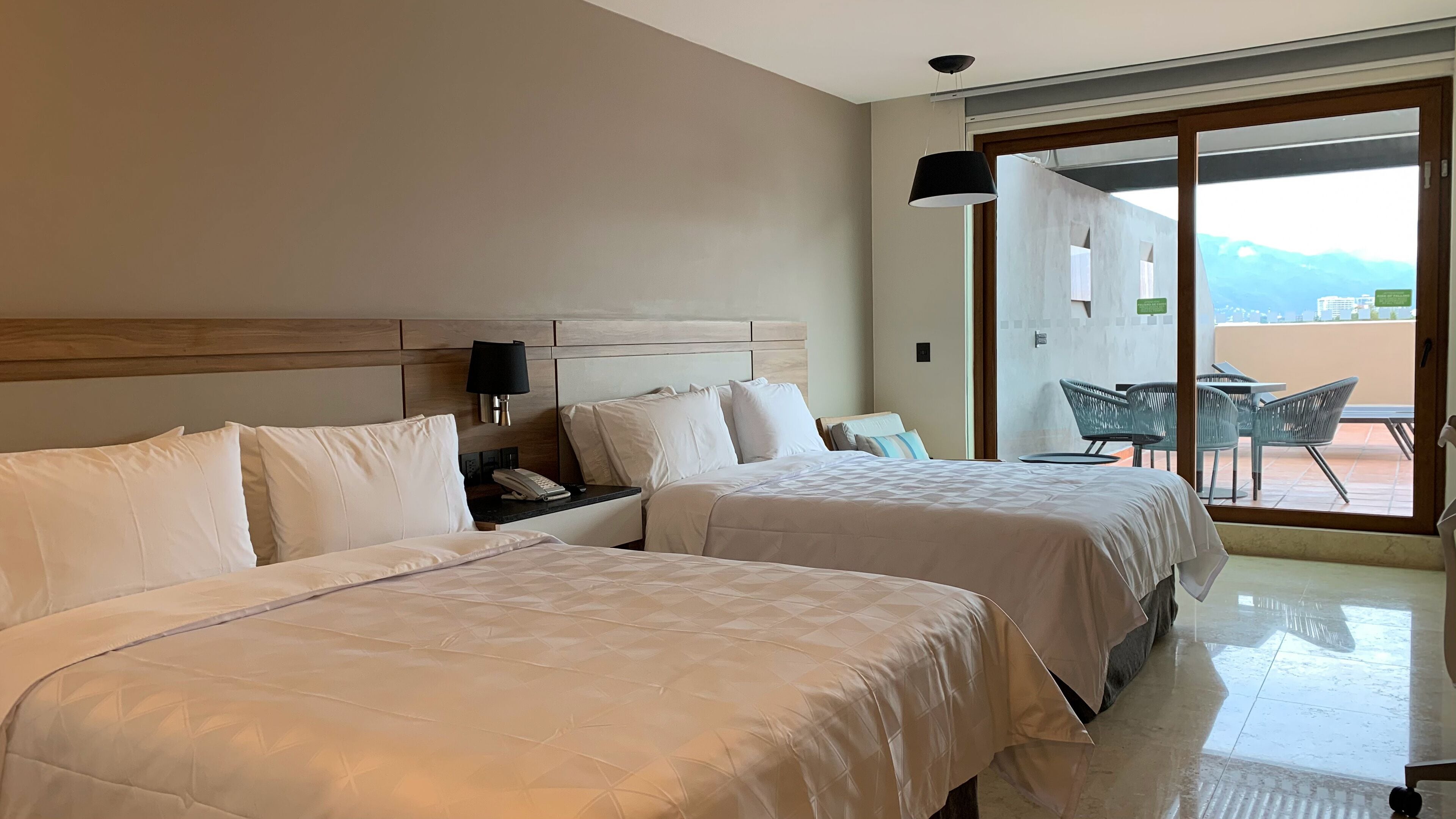 Habitación de hotel con dos camas, una mesita de noche, una lámpara y vistas al exterior a través de una puerta corrediza.