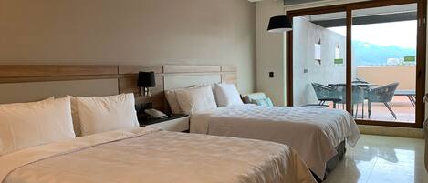 Habitación de hotel con dos camas, una mesita de noche, una lámpara y vistas al exterior a través de una puerta corrediza.