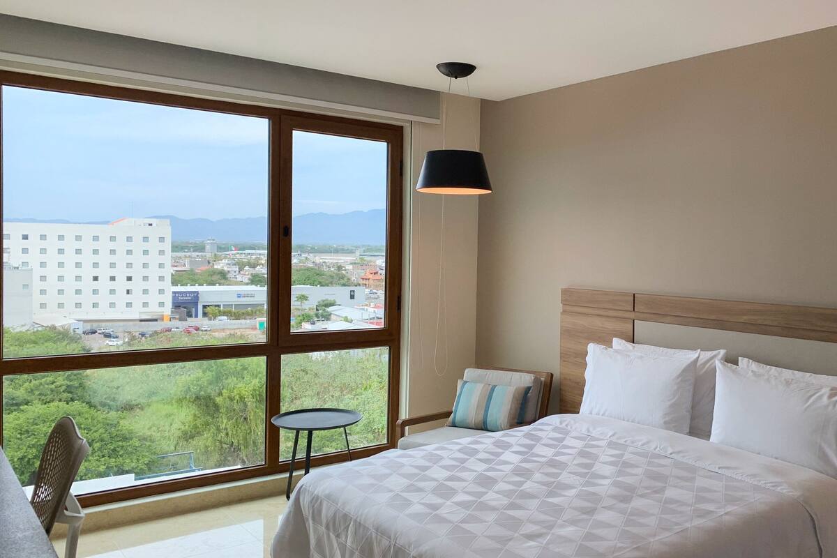 Une chambre d’hôtel dotée d’une grande fenêtre donnant sur la ville, d’un lit avec une literie blanche, d’une petite table et d’une lampe de chevet.