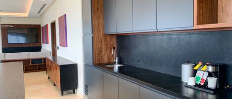 A modern kitchen with grey cabinets, a black countertop, and a built-in oven. There is a wooden cabinet above the counter and a large flat-screen TV mounted on the wall.
