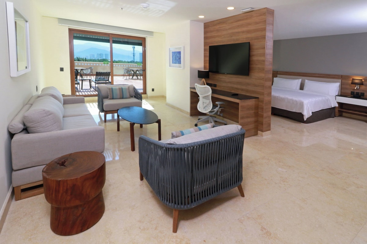A hotel room with a sofa, armchair, TV, bed, and a balcony with chairs.