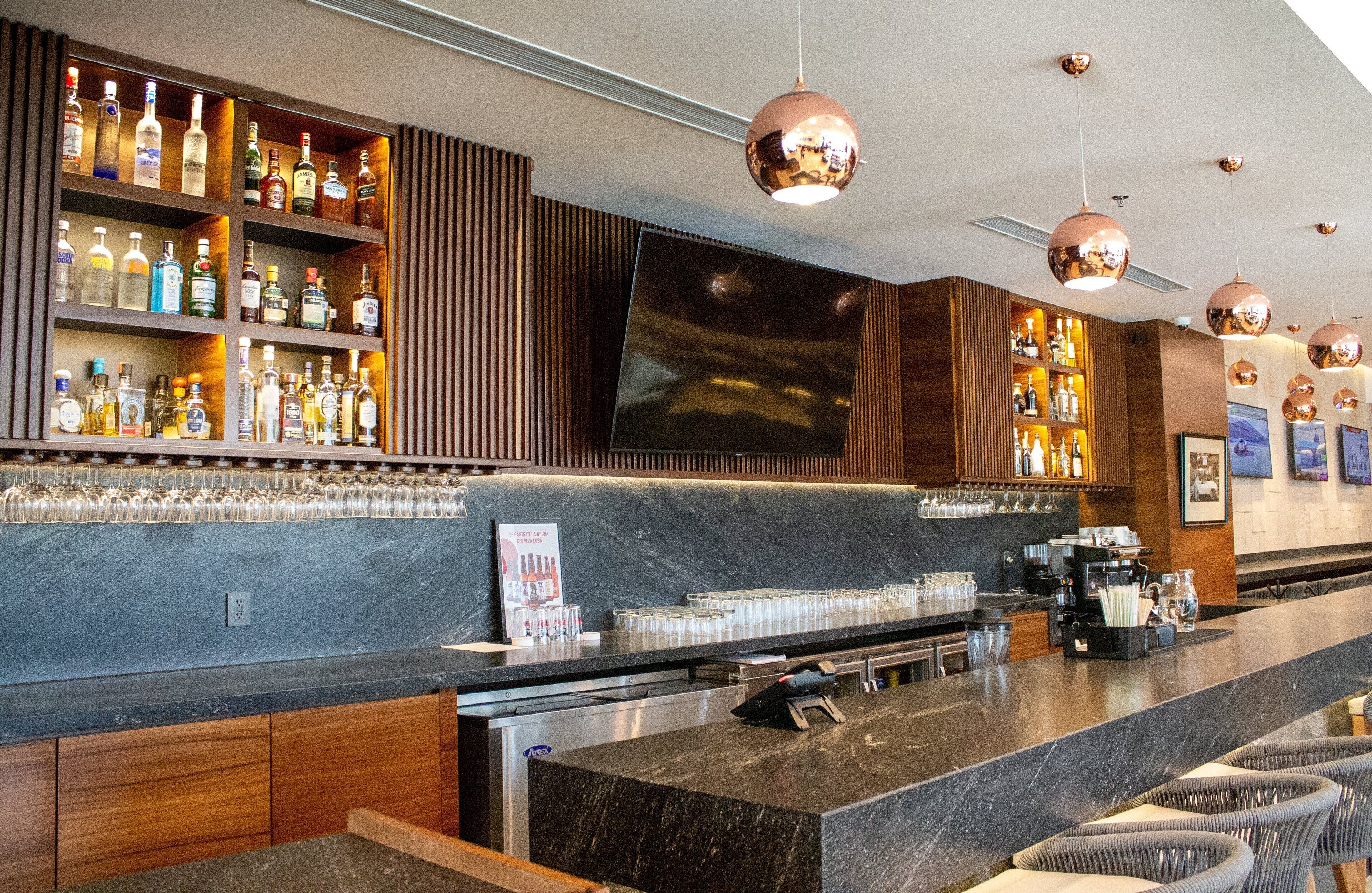 Una zona de bar con una barra larga, estantes con botellas, vasos y una pantalla de televisión grande.