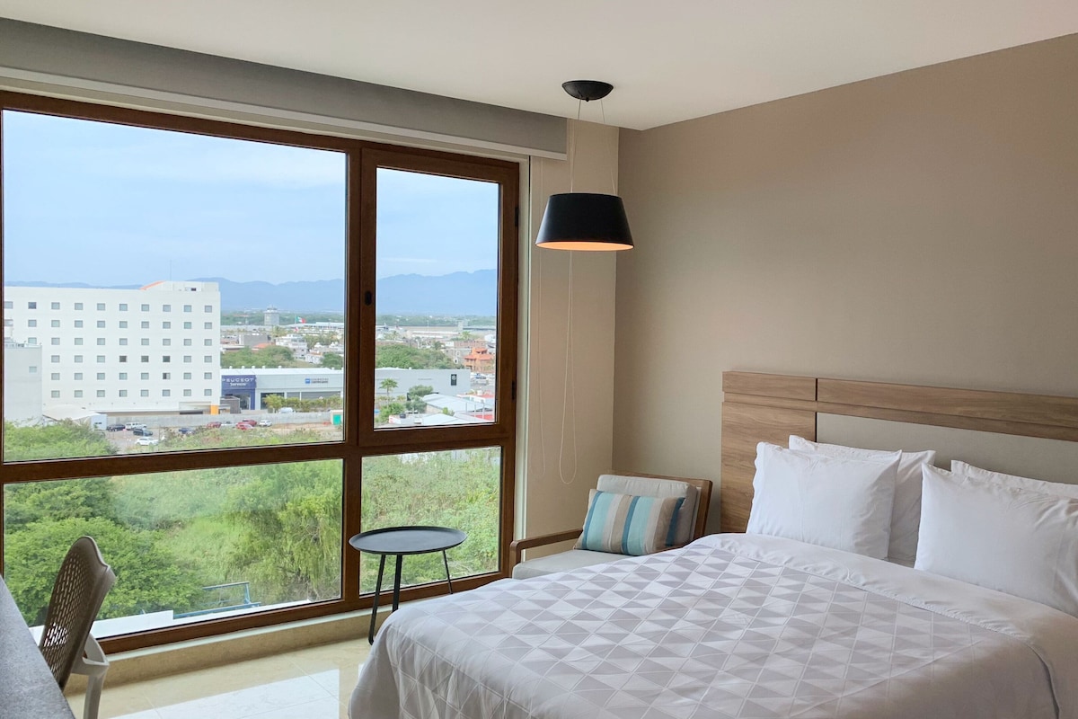 A hotel room with a large window, a bed with white bedding, a bedside table with a phone, and a view of a cityscape.