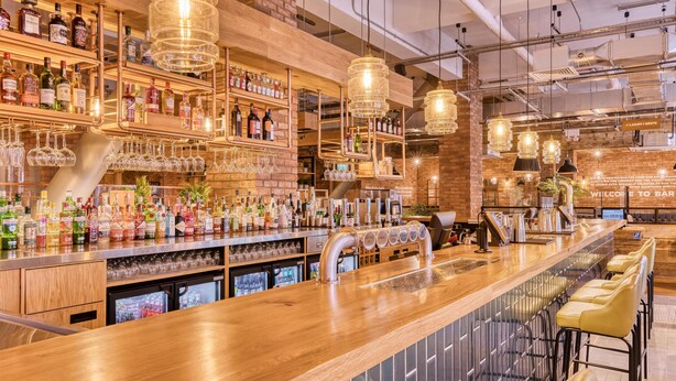 A bar with wooden shelves stocked with bottles and glasses, a long counter with stools, and hanging pendant lights. - Premier Inn Glasgow Cc St Enoch Square (Glasgow)
