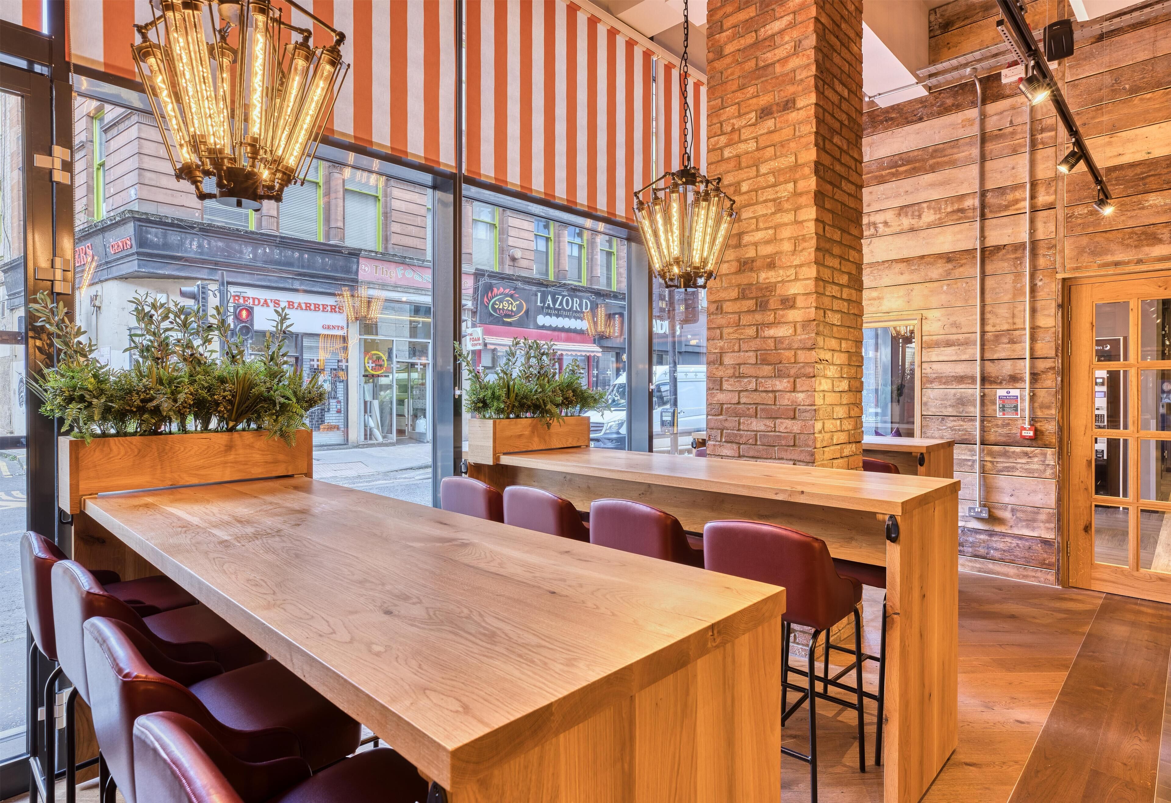 A restaurant interior with wooden tables and chairs, potted plants, and a view of the street outside.