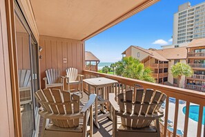 A balcony with Adirondack chairs, a table, and a view of the ocean and hotel buildings.