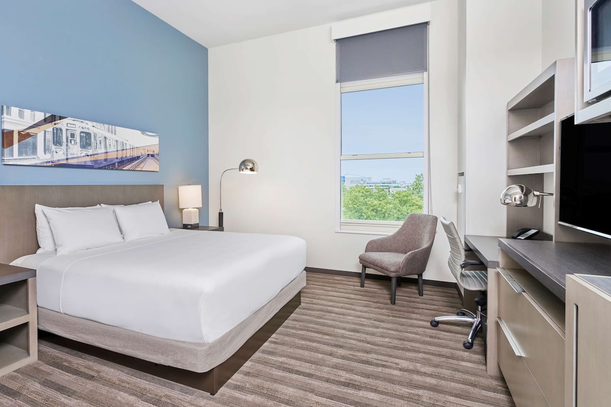 Ein Hotelzimmer mit einem großen Bett, einem Schreibtisch mit Stuhl, einem Fernseher und einem Fenster mit Blick auf die Stadt.