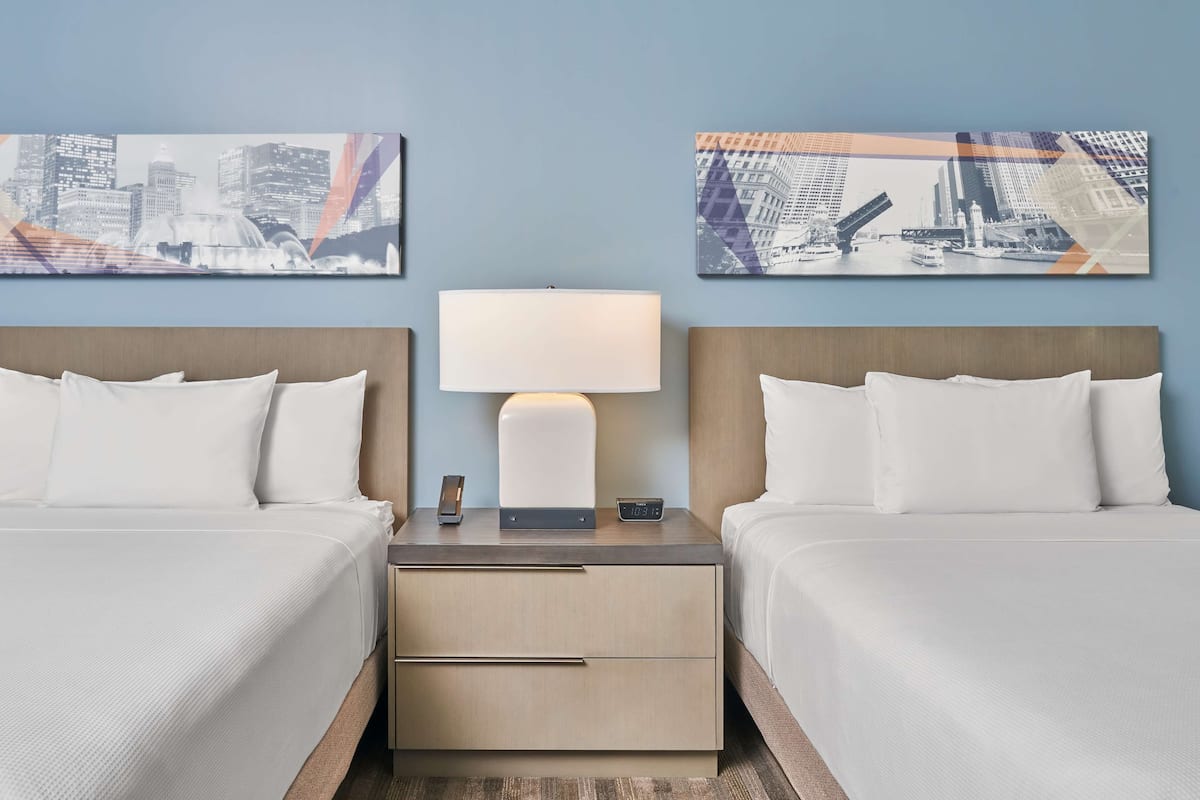 Ein Hotelzimmer mit zwei Betten, einem Nachttisch mit Lampe und zwei Stadtbildern an der Wand.