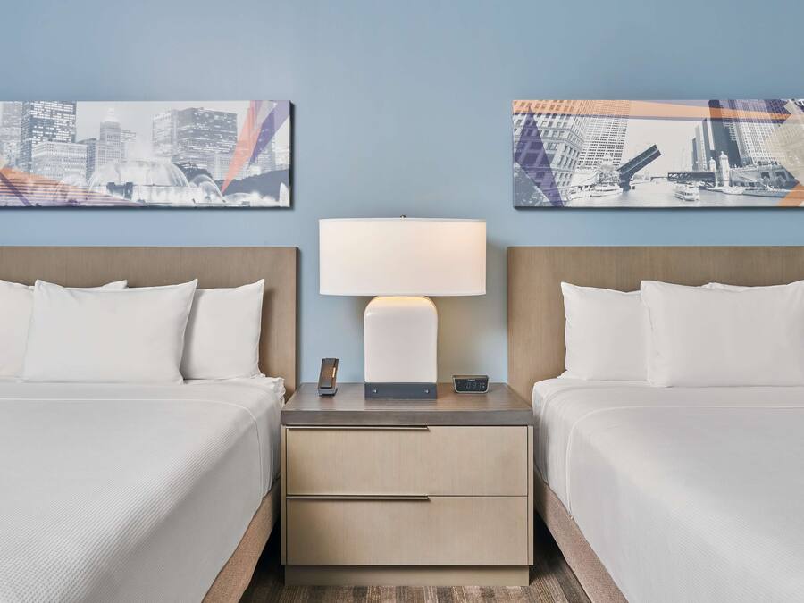A hotel room with two beds, a nightstand with a lamp, and two cityscape paintings on the wall.