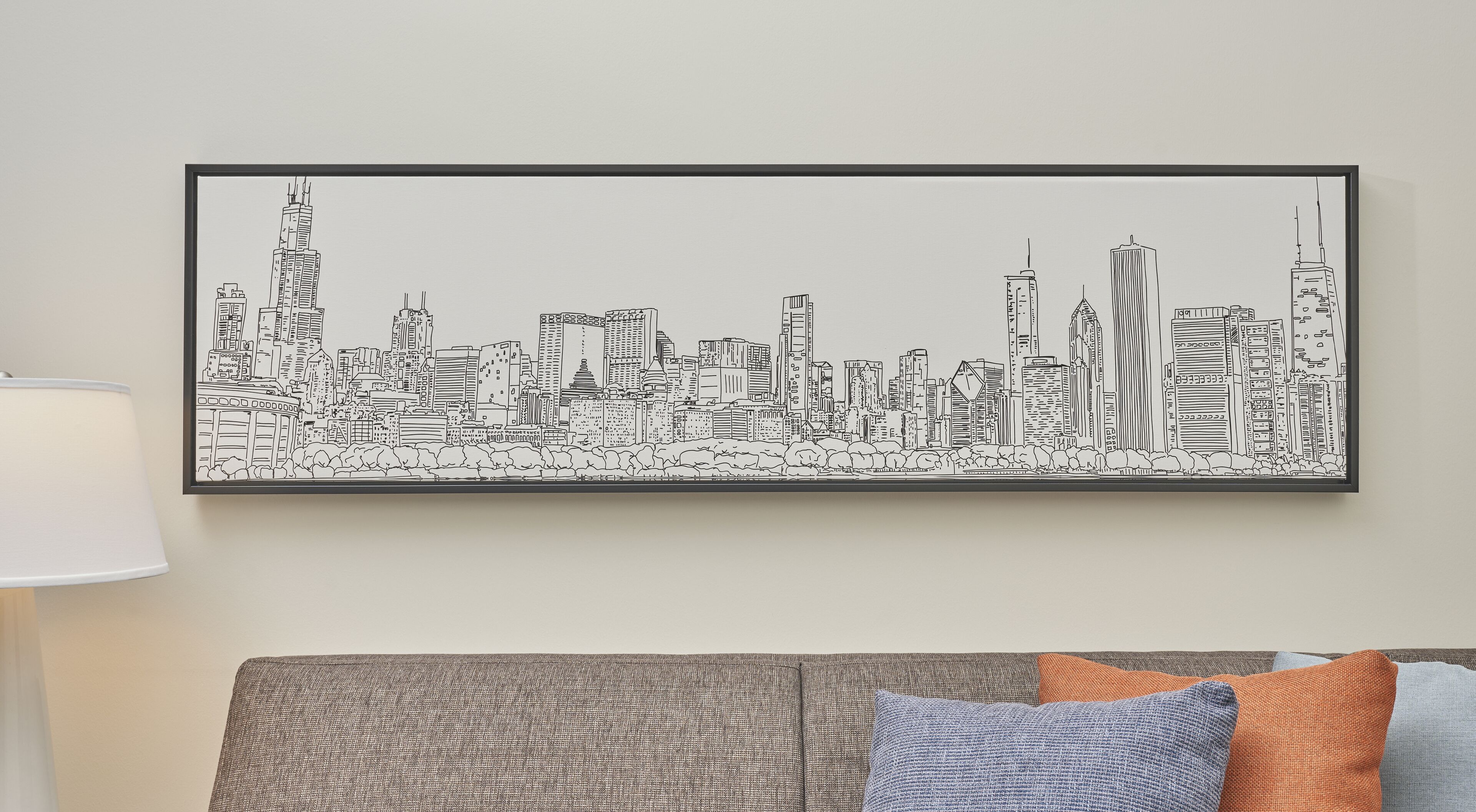 Une œuvre d’art représentant un paysage urbain, encadrée et accrochée au mur au-dessus d’un canapé gris agrémenté de coussins à motifs.