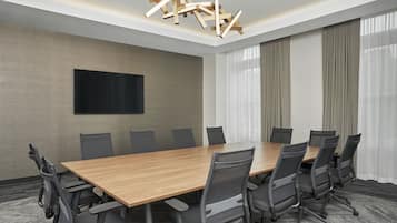 A conference room with a long wooden table, black mesh chairs, a flat-screen TV on the wall, and a modern chandelier.