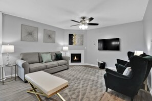 Un salon moderne avec un canapé gris, un fauteuil noir, une cheminée et un ventilateur de plafond.