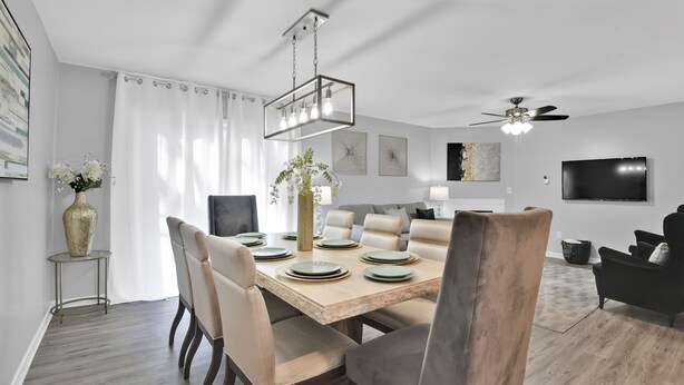 A modern dining area with a wooden table set for six, surrounded by beige chairs. A hanging light fixture and a ceiling fan are visible.