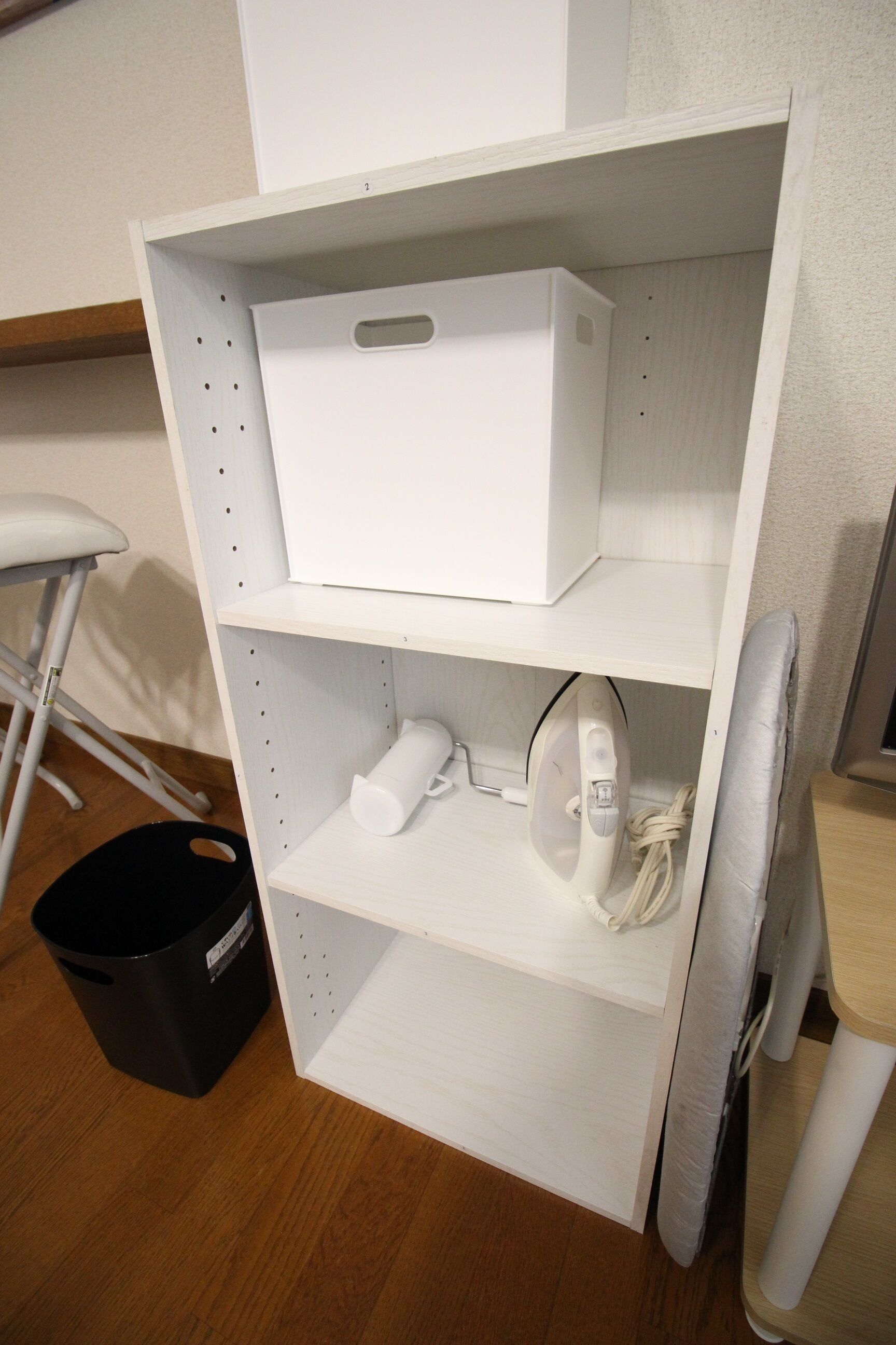 A white shelving unit with a white box on the top shelf, a rolled-up white item on the middle shelf, and an iron on the bottom shelf. A black trash bin is visible to the left.