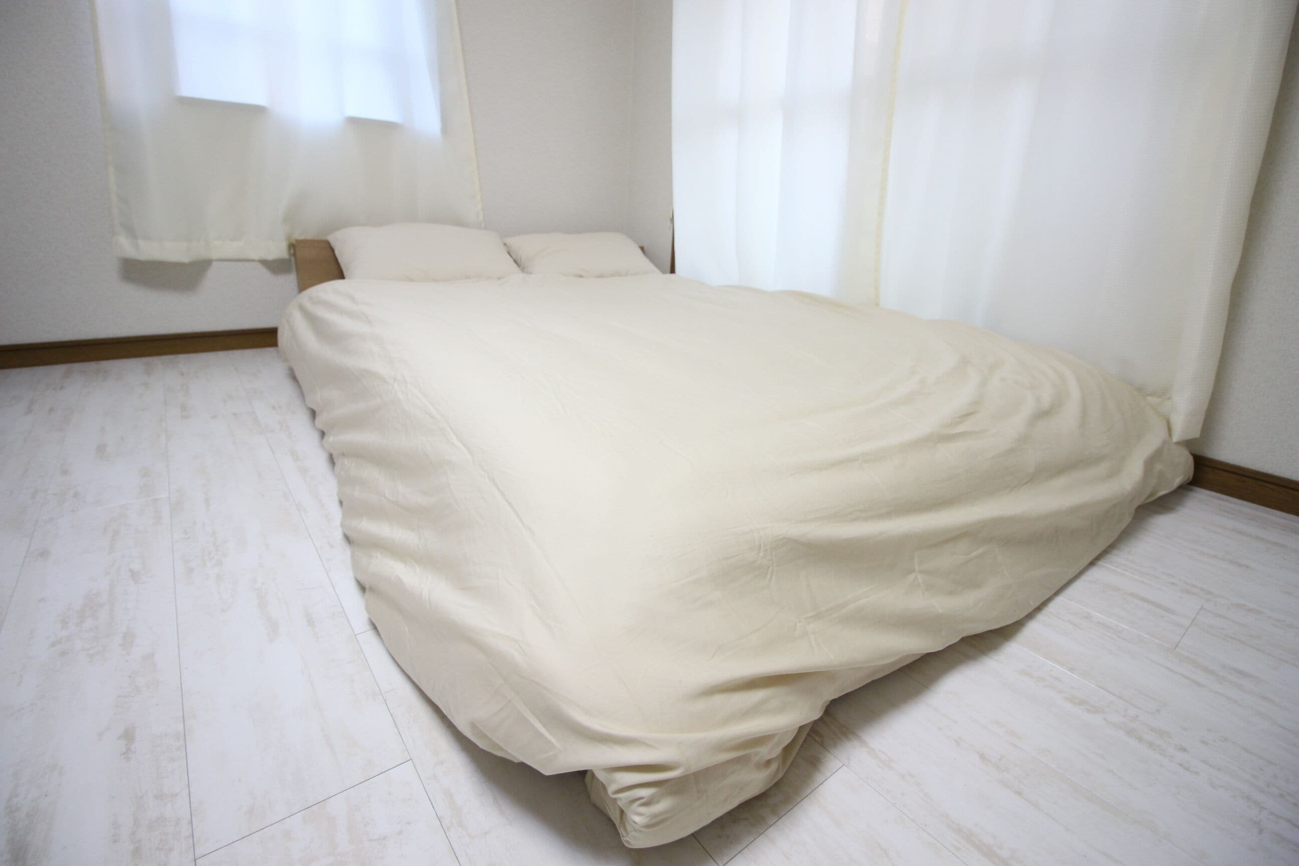 Quarto com cama, cortinas brancas e chão de madeira clara.