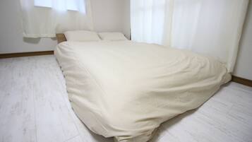Quarto com cama, cortinas brancas e chão de madeira clara.